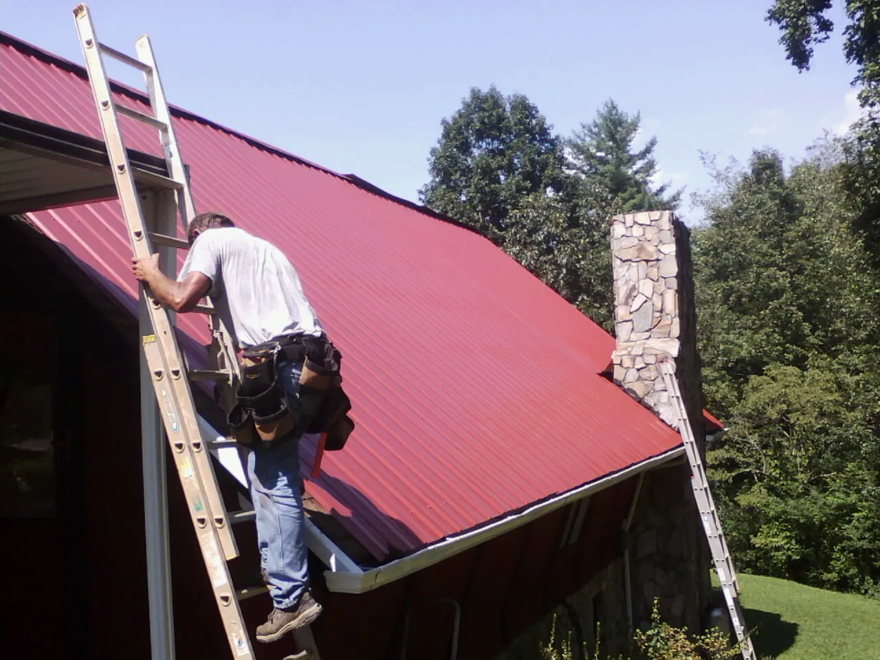 Roofer on a ladder beside a red metal roof and stone chimney — Soffit & Fascia Repair by Roofing Brothers in Airville